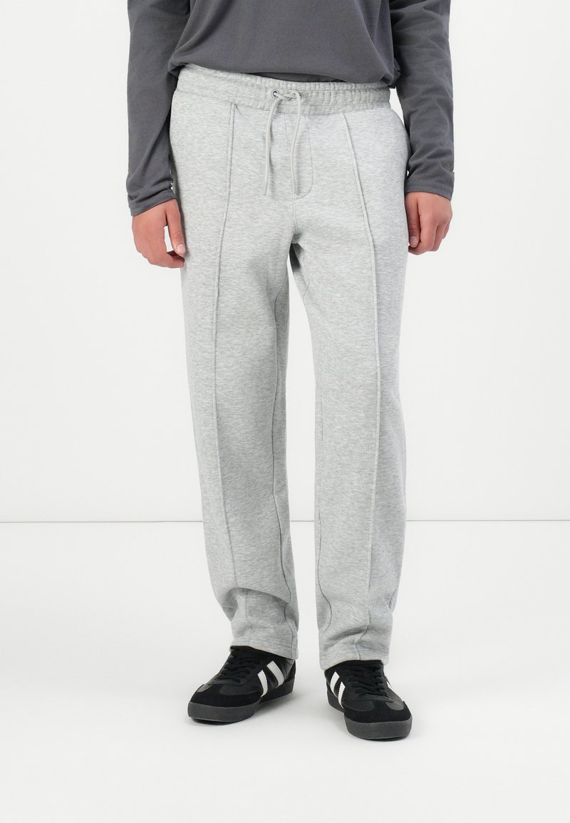 Pantalones de chándal gris claro con cintura ajustable, detalles en las costuras laterales y un corte relajado, combinados con zapatillas negras con rayas blancas.
