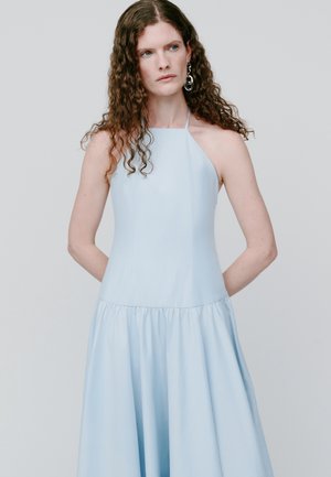 Femme aux longs cheveux bouclés portant une robe sans manches bleu pâle à col halter et de grandes boucles d'oreilles en argent, debout devant un fond clair uni.