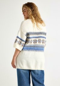 Maglione bianco in lana con motivo geometrico blu e marrone, maniche corte, vestibilità ampia e scollatura tonda, abbinato a jeans in denim blu.