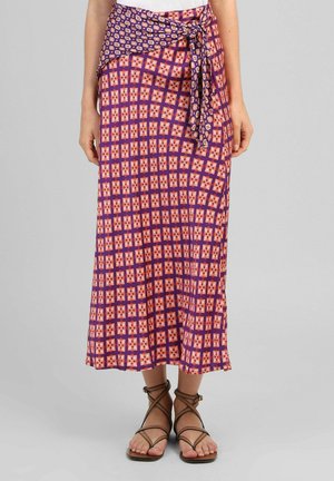 Jupe portefeuille à motifs violets et rouges nouée à la taille, portée avec des sandales plates marron à brides, visible de la taille aux pieds sur fond gris.