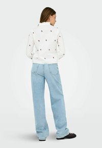 Donna di spalle che indossa una blusa bianca a maniche lunghe con piccoli motivi floreali rossi e jeans a gamba larga blu chiaro con scarpe nere.