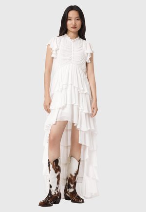 Frau trägt ein weißes mehrlagiges Kleid mit Stufen und asymmetrischem Saum, kombiniert mit kniehohen braun-weißen Cowboystiefeln mit Muster, vor grauem Hintergrund.