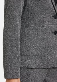 Szary żakiet o fakturowanej strukturze, wykonany z tkaniny plecionej, z klapami w kształcie notcha, dwoma guzikami oraz funkcjonalną kieszenią na piersi. Subtelny wzór w jodełkę.