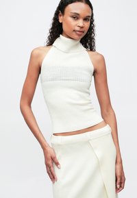 Ungdommelig kvinne med krøllete hår iført ermeløs hvit ribbestrikket turtleneck og matchende skjørt, poserende med hånden på hoften.