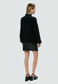 Schwarzer Strickpullover mit strukturierten Puffärmeln, kombiniert mit einem schwarzen Leder-Minirock und klobigen Absatzschuhen, von hinten betrachtet.
