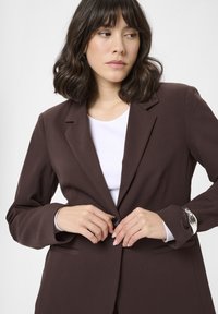 Bruine op maat gemaakte blazer met een enkele knoopsluiting, notch-lapels en zijzakken, gedragen over een wit shirt. Zachte, gestructureerde stof.