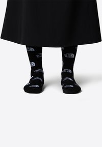 Svarta strumpor med vit text och logotyper som har "THE NORTH FACE" i ett upprepat mönster. Strumporna har en standardform och -tjocklek.