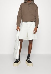 Man wearing brown hoodie and white shorts, holding white tote bag, black socks, and black platform slip-on shoes against white background.