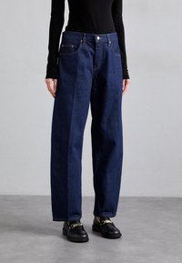 Tmavě modré široké džíny s vysokým pasem, vybavené knoflíkovým zapínáním, kapsami a klasickou dením texturou. Spárovány s černými botami.