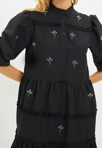 Abito nero con maniche a palloncino corte e chiusura frontale con bottoni. Presenta ricami floreali blu chiaro e volants a strati. Tessuto morbido.