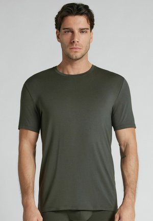 Joven con cabello corto y oscuro que lleva una camiseta lisa de color verde oscuro con mangas cortas, de pie frente a un fondo gris claro.