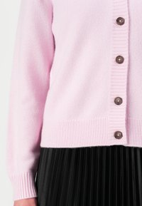 Cárdigan de un suave tejido en color rosa claro, con dobladillos y puños acanalados, abrochado con botones marrón oscuro, combinado con una falda negra con pliegues.