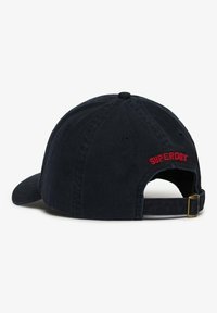 Gorra de béisbol azul marino confeccionada en algodón, con visera curva, correa ajustable y logo bordado en rojo de "SUPERDRY" en la parte trasera.