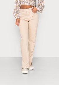 Jeans retos bege claro com uma bainha ligeiramente desfiada e aberturas laterais, feitos de tecido de ganga. Combinados com sapatilhas brancas.