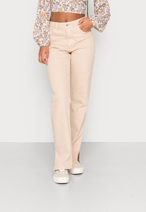 Jeans retos bege claro com uma bainha ligeiramente desfiada e aberturas laterais, feitos de tecido de ganga. Combinados com sapatilhas brancas.