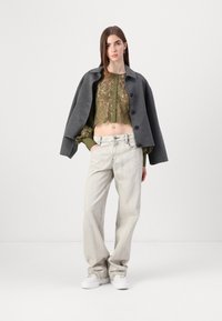 Manteau gris court porté sur une blouse verte brodée, associé à un jean large gris clair et des baskets blanches. Fond uni.