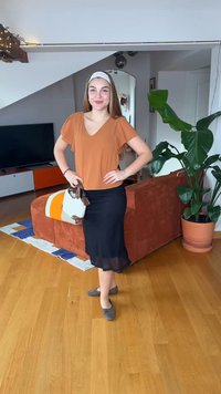 Νεαρή γυναίκα με ανοιχτό καστανά μαλλιά, που φορά πορτοκαλί μπλούζα και μαύρη φούστα, στέκεται στο σαλόνι κρατώντας μια πολύχρωμη τσάντα χειρός, χαμογελώντας.