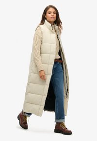 Gilet lungo beige imbottito con colletto alto, design trapuntato, tasche laterali e zip. Indossato sopra un maglione crema a trecce e jeans blu.