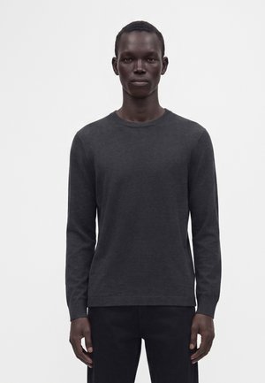 Homme portant un pull à manches longues gris foncé et un pantalon noir, debout devant un fond clair uni.