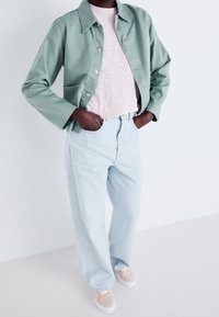 Jasnozielona bawełniana kurtka, różowa tkaninowa koszulka, jasnoniebieskie dżinsy, beżowe sneakersy z różowymi akcentami. Model stoi z rękami w kieszeniach.