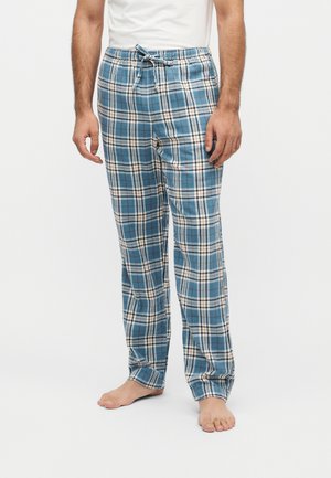 Homme portant un pantalon de pyjama à cordon bleu et blanc à carreaux, pieds nus sur un fond blanc uni.