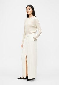 Naine seisab, kandes kreemjat soonikutega kampsunit, mis on toppitud kõrge vöökohaga kreemjate laia säärega pükstesse, millel on ees hõlm, ning musti teravate ninaga kingi.