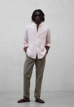 Hombre con rastas y gafas de sol, de pie con las manos en los bolsillos, vistiendo una camisa de botones rosa claro, pantalones caqui y chanclas negras.