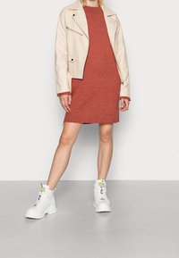 Beige lädermotorcykeljacka över en rostfärgad stickad klänning, parat med vita chunky stövlar med gula detaljer och svart text.