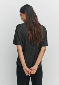 Schwarzes T-Shirt mit kurzen Ärmeln und lockerer Passform sowie einer subtilen Textur. Getragen mit schwarzen, weiten Hosen. Die Rückansicht zeigt lange Haare.