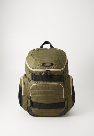 Mochila verde oliva con textura acolchada, herrajes negros, correas ajustables, bolsillo frontal con ribete beige y bolsillos de malla laterales.