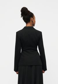 Veste noire cintrée avec manches longues, taille ajustée et col pointu, associée à une jupe plissée noire. Texture de tissu lisse.