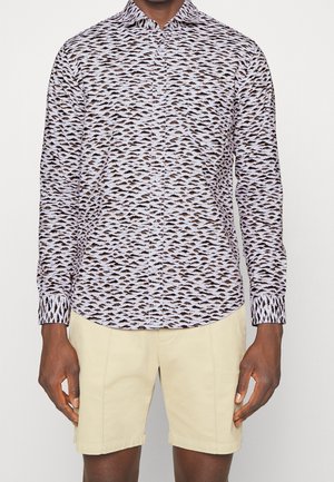 Uomo che indossa una camicia a maniche lunghe con un piccolo motivo montano e pantaloncini beige fino al ginocchio, in piedi davanti a uno sfondo bianco.