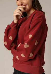 Pull rouge à manches longues avec des broderies en forme de cœur beige le long des bras ; poignets et ourlet côtelés, fabriqué en matériau doux.