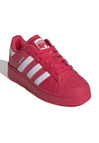 Röd Adidas Superstar-sneaker med vita ränder, texturerad skalm tå och "Superstar"-text på sidan, vinklad för att visa ovansidan och yttre sidan.