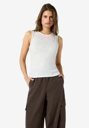 Femme portant un débardeur blanc et un pantalon cargo ample marron foncé, les mains dans les poches, sur un fond blanc uni.