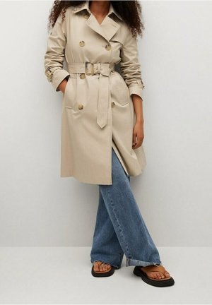 Frau trägt einen beigen, taillierten Trenchcoat, blaue weit geschnittene Jeans und braune offene Sandalen, steht vor einer einfachen Wand.