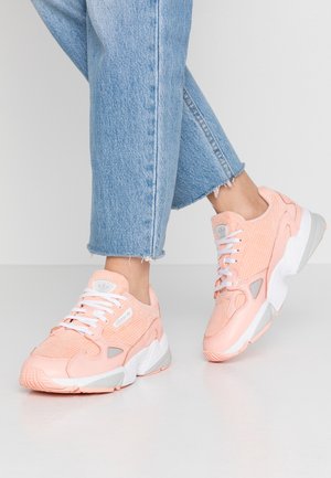 Persoon draagt lichtblauwe spijkerbroek en roze Adidas sneakers met witte zolen, staand op een gladde lichtgrijze vloer.