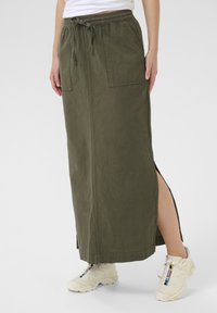 Jupe en coton longue, de couleur vert olive, avec une taille élastique à cordon de serrage, deux poches avant et des fentes sur les côtés. Associée à des baskets blanches.