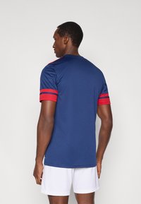 Marineblaues Sporttrikot mit roten Streifen an den Ärmeln. Aus leichtem Stoff, kurzärmelig, mit rundem Halsausschnitt.