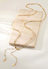 Gold-tone necklace featuring layered chains, faceted beads, and decorative clasps, displayed on a textured beige stone surface.
