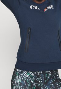 Marineblauwe trui met wit en oranje logo, twee verticale zakken met rits, gedragen met zwarte en groene legging met patroon.