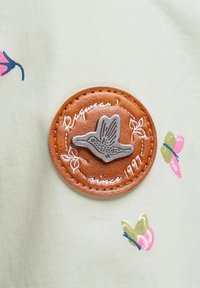 Parche de cuero marrón con un pájaro plateado en relieve y texto manuscrito, cosido a una tela verde claro con patrones florales rosas y verdes.