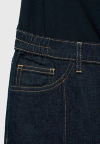 Mörk denimjeans med orange sömmar som visar midjeband, bältesögla och detaljer på bakfickan mot en svart topp.