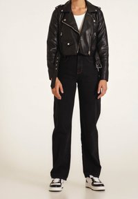 Chaqueta de motorista de cuero negra corta con cremalleras y botones, combinada con jeans negros de cintura alta y zapatillas en blanco y negro. Camiseta blanca lisa debajo.