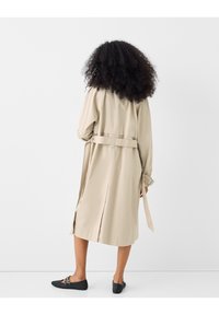 Beige trenchcoat med bälte i midjan, långa ärmar och en vent bak. Bärs med svarta vävda slip-on skor, sedd bakifrån.