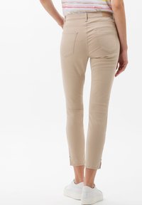 Mujer con pantalones cropped ajustados beige y zapatillas blancas, de pie con una mano en la cadera, vista desde atrás.