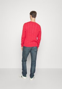 Pull en maille torsadée rouge vif, avec col et poignets côtelés, associé à un jean bleu foncé et des baskets blanches avec des accents verts.