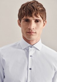 Młody mężczyzna z krótkimi brązowymi włosami, ubrany w białą, zapinaną na guziki koszulę, na jasnym jednolitym tle.