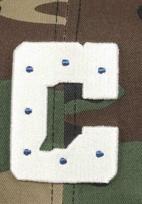 White embroidered letter "C" with blue dots on a camouflage fabric background in green, brown, and tan tones.