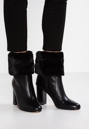 Botines de tacón negro con puños de piel sintética, acabado de cuero suave, puntera cuadrada y tacón grueso. Cuentan con un cierre lateral con cremallera y herrajes decorativos.